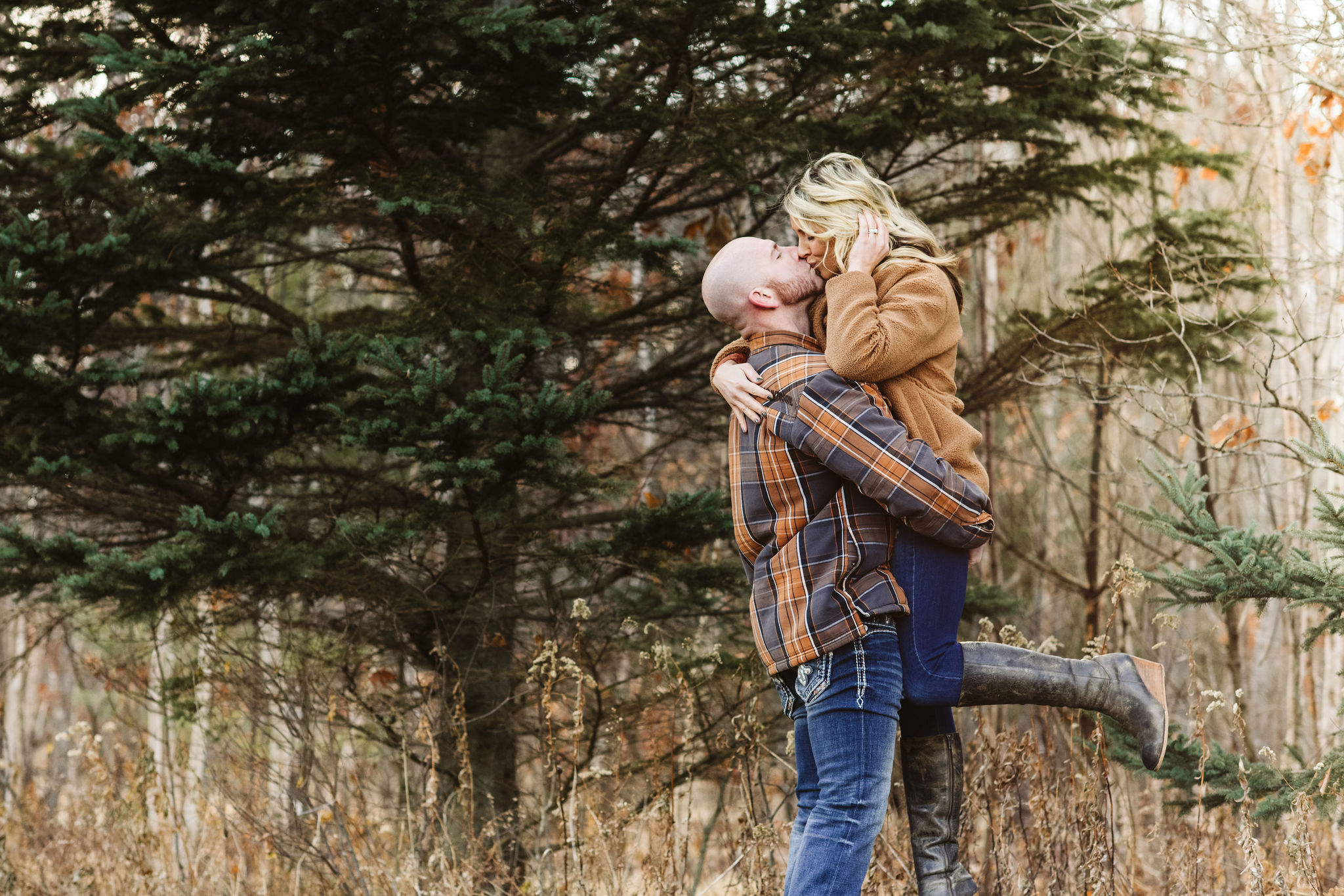 A man lifts his wife up for a kiss during their family photo session. There is a pine tree in the background, and some golden dried grasses around it. They are both wearing jeans and a casual top.