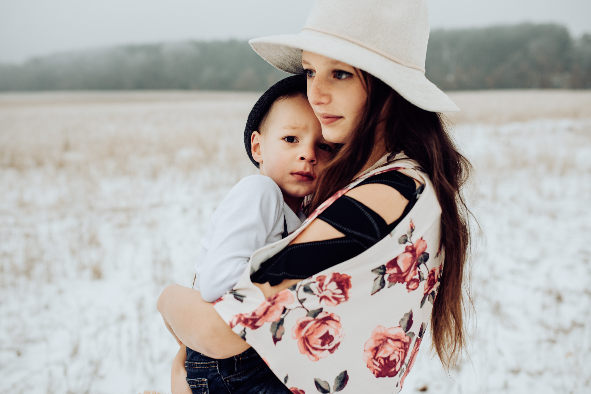 A girl carries her young brother in a snowy meadow during their family photos in the snow. She is wearing a white hat and a floral dress.