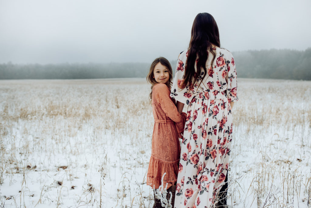 A young girl smiles at the camera during their family photos in the snow. She is wearing a pink dress and standing by her older sister. They stand in a snow covered field.