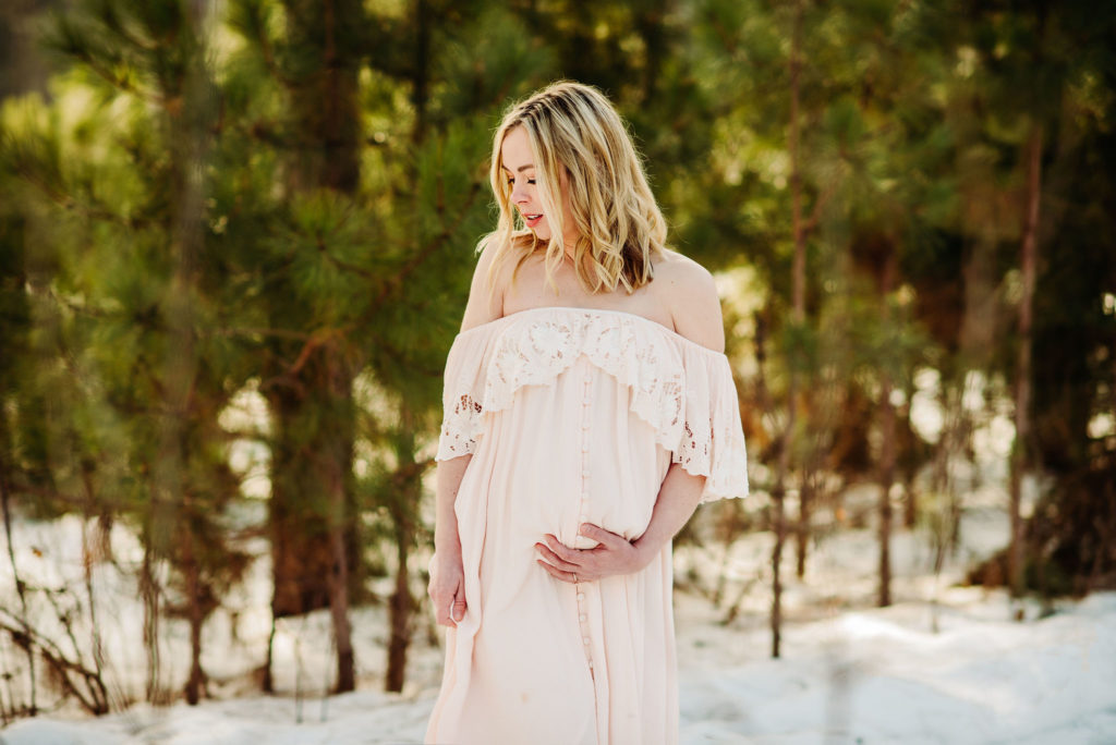 A maternity photo of a woman in an ivory, off the shoulder dress. She is gazing down and to her right. She has her left hand under her belly. With her right hand she holds the long, flowy skirt of the dress. There is snow on the ground and pine trees behind her.