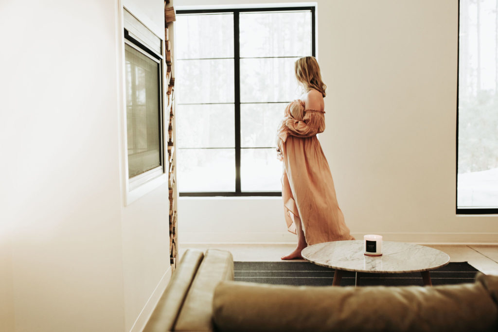 A pregnant woman walks by the window at the Pinewood Wedding and Events venue. She is looking out the window and cradling her belly. She is in a flowy, pink, off the shoulder dress. She gazes out the window.