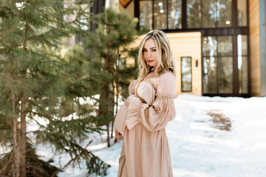 Maternity photo of a blond woman in front of the Pinewood Wedding and Events venue. There is snow on the ground and pine trees to her left. The windows of the wedding venue can be seen behind her. She has her hands lovingly around her belly. She is looking at the camera and grinning.