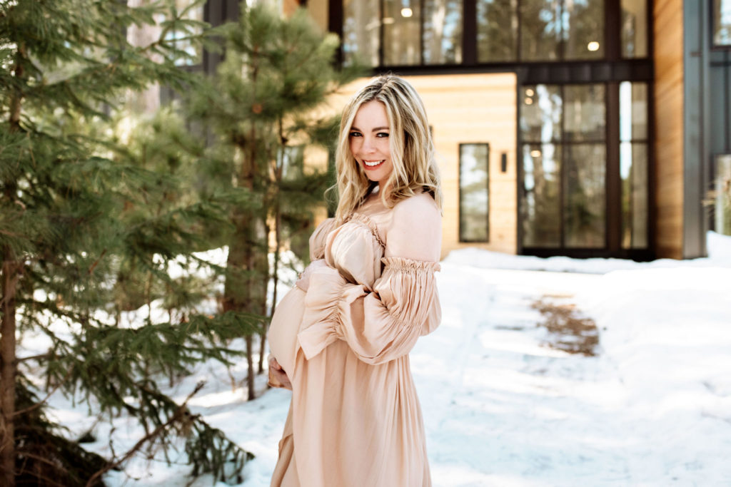 Minnesota maternity shoot of a blond woman in front of the Pinewood Wedding and Events venue. She is smiling and looking at the camera. There is snow on the ground and pine trees to her left. The windows of the wedding venue can be seen behind her. She has her hands lovingly around her belly.