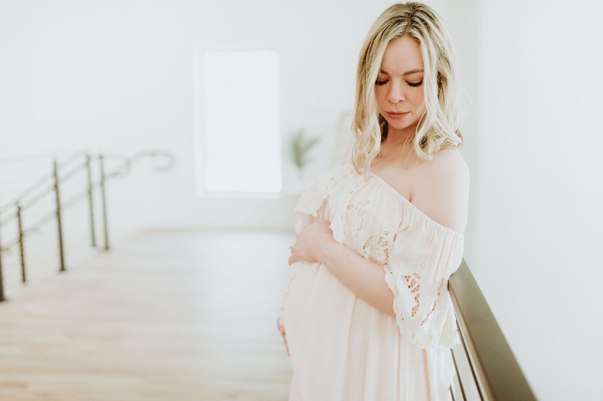 A pregnant woman looks down over her shoulder while holding her belly. She is in an all white space with window light coming in from behind her. The space is Pinewood Weddings and Events in Cambridge Minnesota.
