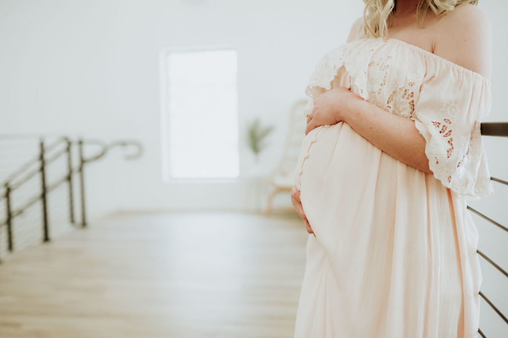 A close up photo of a pregnant woman cradling her belly. She is in an all white space with window light coming in from behind her. The space is Pinewood Weddings and Events in Cambridge Minnesota.