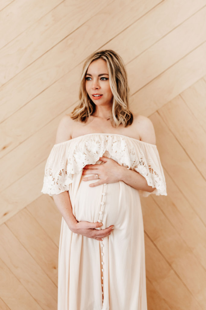 A pregnant woman is holding her belly. She is gazing to the side with a peaceful and eager expression. She is wearing an ivory Fillyboo dress. The dress is off the shoulder with lace around the top and buttons down the middle. She stands in front of a wood panel door.