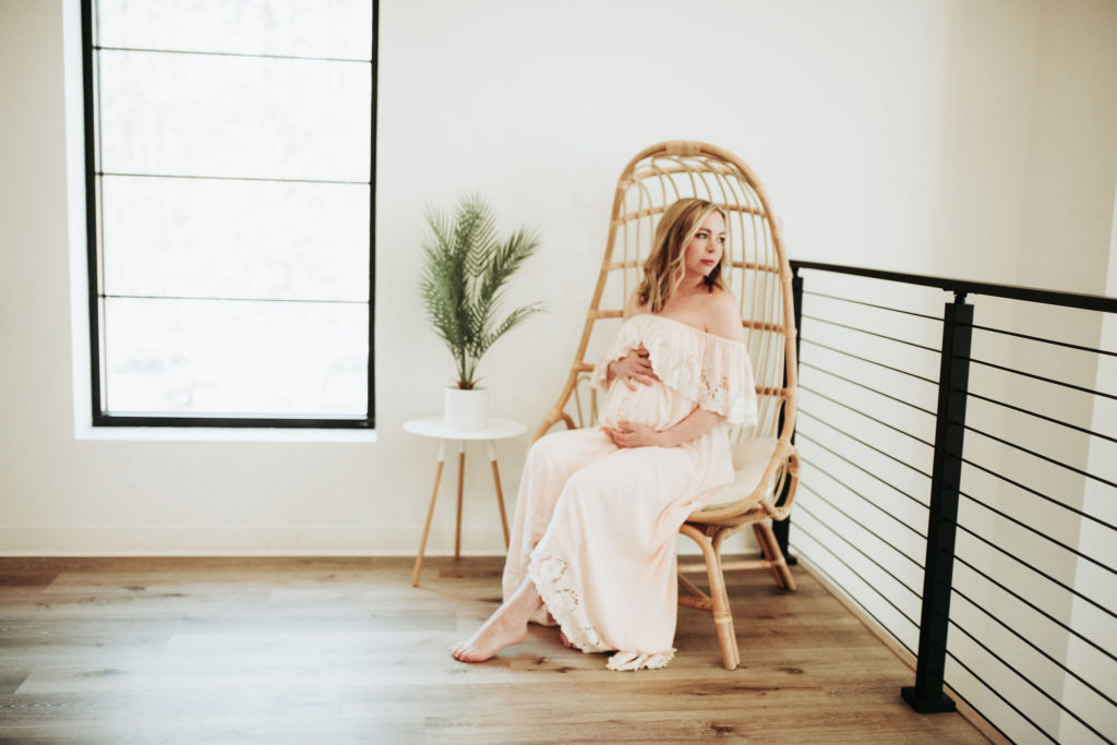 A maternity photo. A pregnant woman sits in a wicker high backed chair. She is looking over her shoulder into the distance while cradling her belly. Minnesota maternity shoot held at the Pinewood Wedding and Event venue. The decor is minimal. The walls are white and railing is black.
