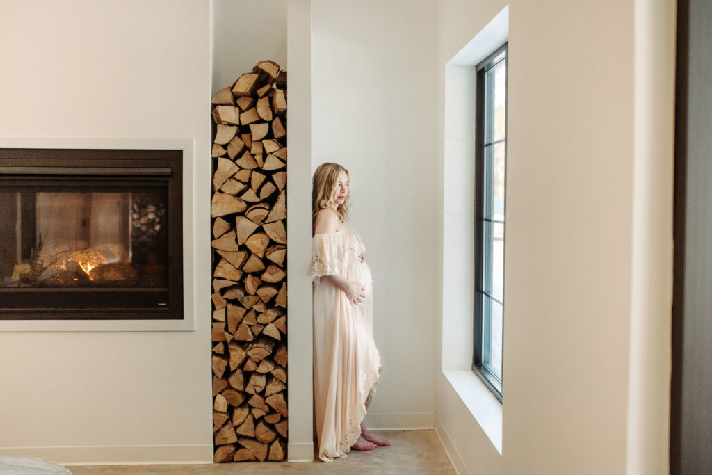 Pregnancy photo. A woman stands by a fireplace. There is a decorative wall holding firewood behind her. She is pregnant and is lovingly holding her belly. She is wearing a long, ivory Fillyboo dress. She is looking out the window with her back against the wall. She is at the Pinewood Wedding and Events venue. The walls around her are white. The trim and fireplace are black.