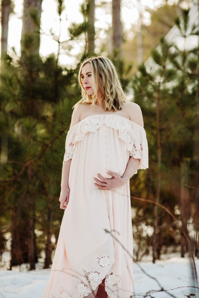 A maternity photo of a woman in an ivory, off the shoulder Fillyboo dress. She is looking off to her right. She has her left hand lovingly under her belly. With her right hand she holds the long, flowy skirt of the dress. There is lace at the bottom of the dress and around the off the shoulder neckline. There are small ivory buttons down the full length of the dress. 