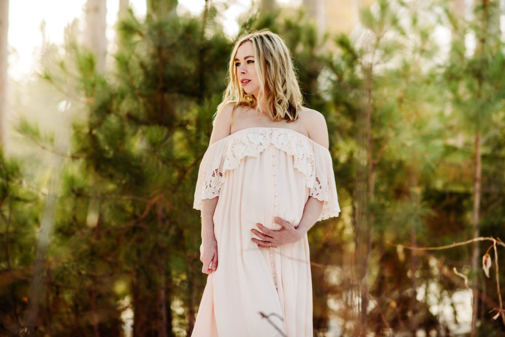 A maternity photo of a woman in an ivory, off the shoulder dress. She is gazing out and to the right. She has her left hand under her belly. With her right hand she holds the long, flowy skirt of the dress. There are pine trees behind her. There is snow on the ground behind the pine trees.