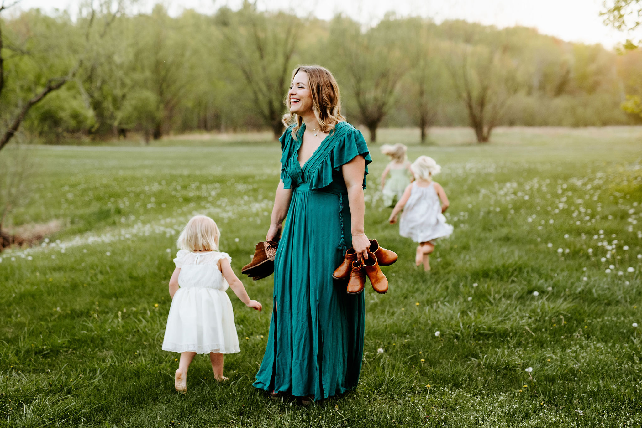 A mother stands in a field full of white dandelion flowers that have gone to seed. She is wearing a turquoise dress. She laughs while she holds several pairs of shoes. Three young girls are running away from her, through the flowers. The sun is setting in the background.